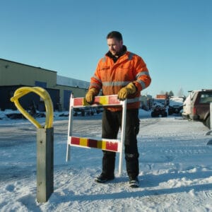 Arbetare i varselkläder bär en hopfälld LA-grind, arbetsgrind i aluminium med röd-gula reflexer på en snöig arbetsplats.