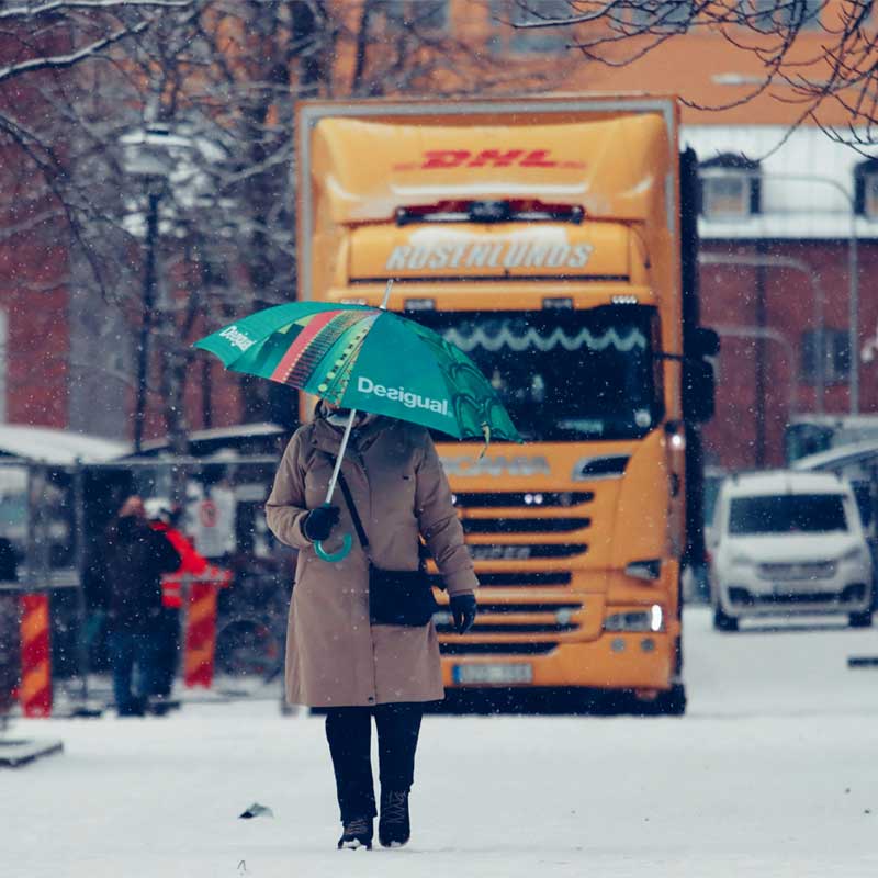 Farthinder för ökad trafiksäkerhet vid tung trafik Äldre dam med paraply går genom en snöig svensk väg medan ett tungt fordon passerar i bakgrunden. Bilden illustrerar vikten av farthinder och trafiksäkerhet vid genomfartstrafik i bostadsområden.