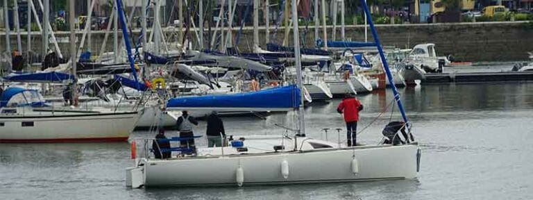 Segelbåtar ligger förtöjda i hamn vid en båtklubb där farthinder och vägbommar används för att öka säkerheten i området.