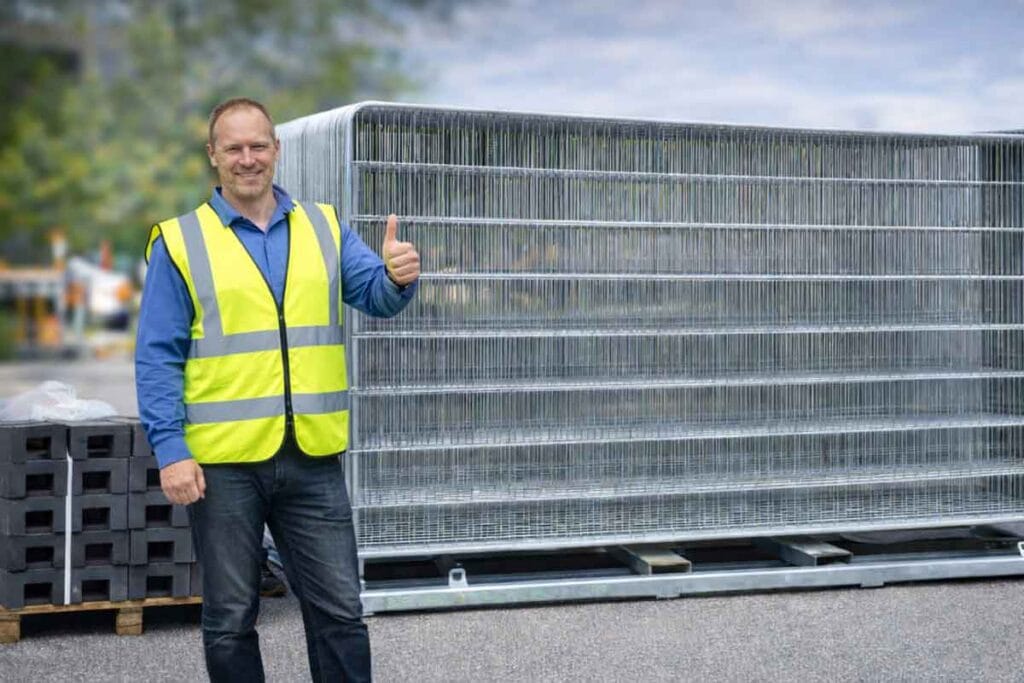 Byggstängsel i galvaniserat stål med man i reflexväst som ger tummen upp på byggarbetsplats.