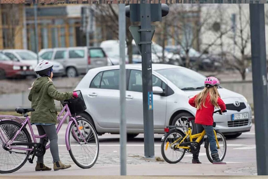 Två barn med hjälmar står med cyklar vid en gata och väntar på att korsa vägen medan bilar passerar.
